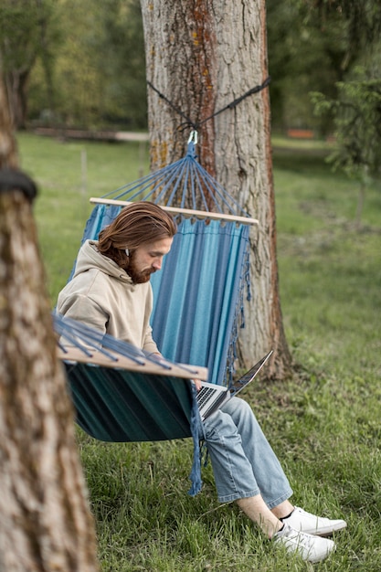 Free photo man in nature sitting in hammock and working on laptop