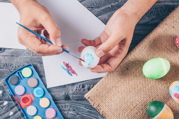 man painting an egg with easter eggs on sack and paint on dark wooden background