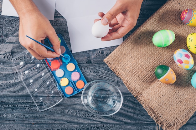 man painting an egg with easter eggs on sack and paint top view on dark wooden background