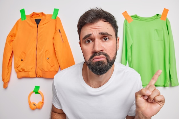 Free photo man points at clothes plastered to wall feels sad poses around orange jacket green jumper and headphones makes choice promots something