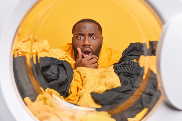 Free photo man poses from inside of washer puts laundry into washing machine has stunned expression dark skin does housework washes clothes at home