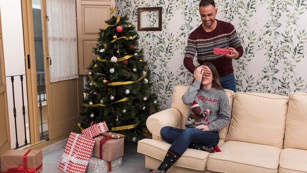 Man presenting gift box and closing eyes to woman on settee near Christmas tree