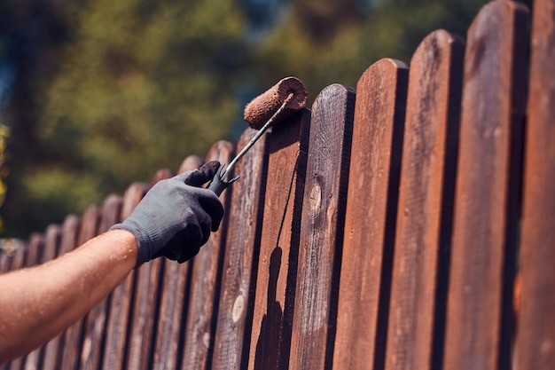 Man in protective gloves is painting wooden fence in bright summer day.