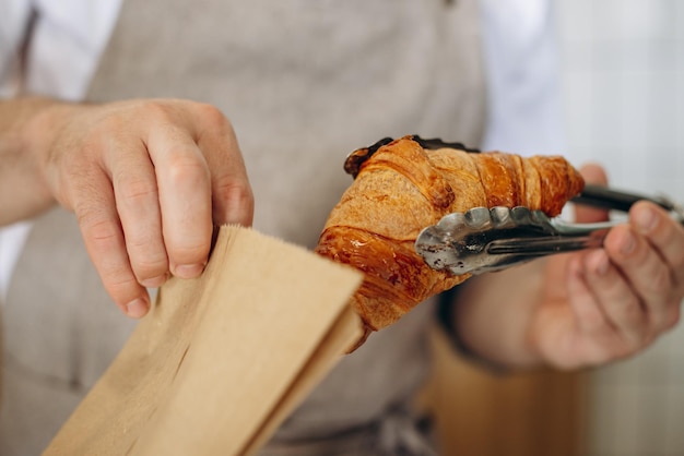 Man puts croissant into a paper bag using tongs