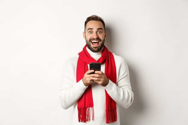 Man reading amazing promo offer in internet, holding smartphone and looking surprised, standing in winter sweater against white background.