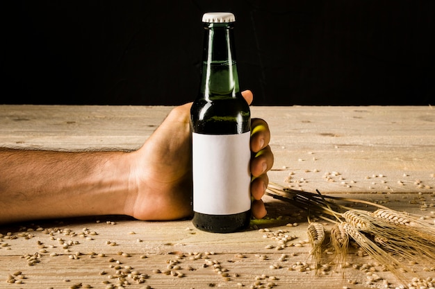 Man's hand holding beer bottle with ears of wheat on wooden surface