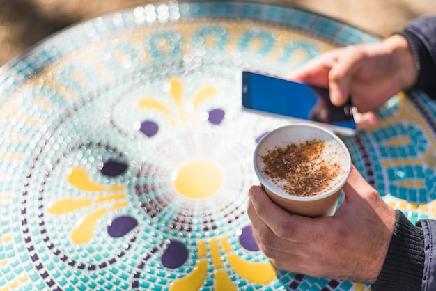 Free Photo man's hand holding cups of cappuccino sprinkled with powdered chocolate using mobile phone