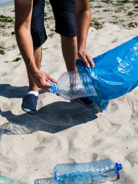 Man's hand putting plastic bottle in blue trash bag on beach