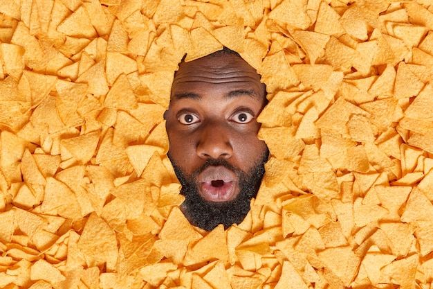 Free photo man shows face through heap of mexican chips keeps mouth opened from great wonder has thick beard cannot believe his eyes eats unhealthy snack