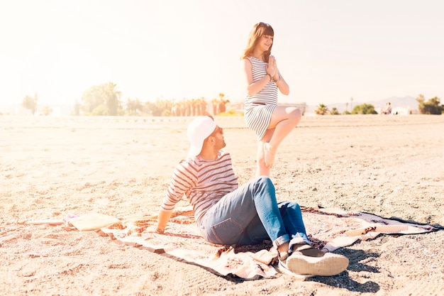 Man sitting on blanket looking to her girlfriend doing yoga at beach