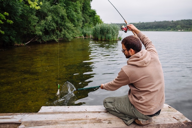 Free photo man sitting on pier catching fish with fishing rod