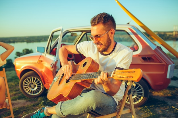 Free photo man sitting and resting on the beach playing guitar on a summer day near river. vacation, travel, summer concept.