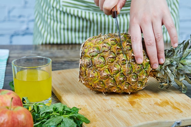 Man slicong a fresh poneapple on wooden cutting board next to apples and a glass of juice.