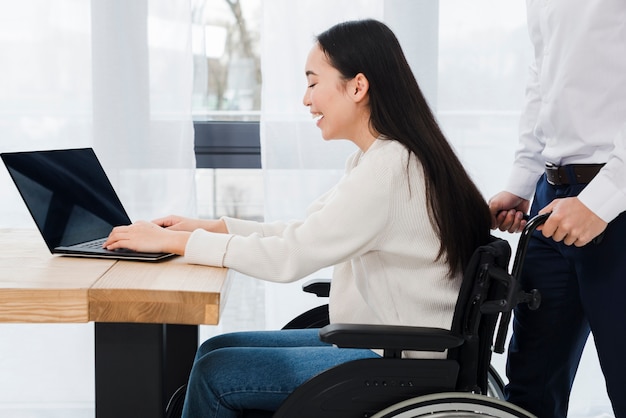 Man standing behind the smiling disabled woman sitting on wheelchair using laptop