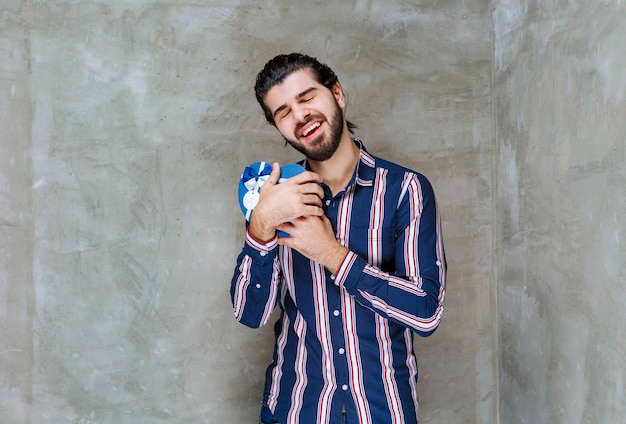 Man in striped shirt holding a blue heart shape gift box and hugging it as feels very happy