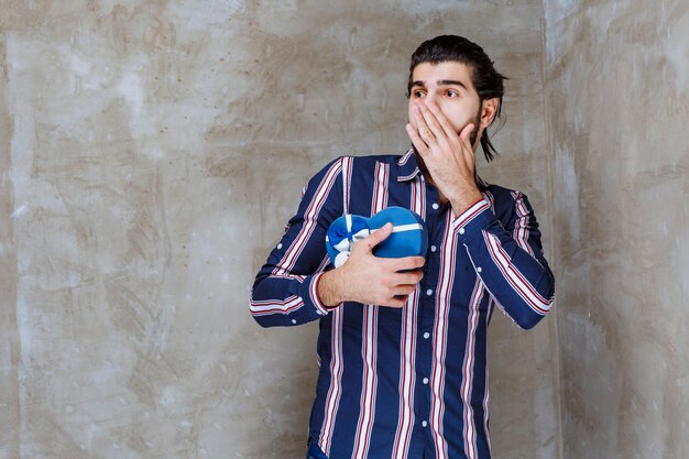 Man in striped shirt holding a blue heart shape gift box and looks thrilled and scared