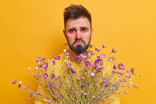 Free Photo  man suffers from rhinitis holds bouquet of flowers reacts on allergens has red watery eyes has unhappy expression poses on yellow 