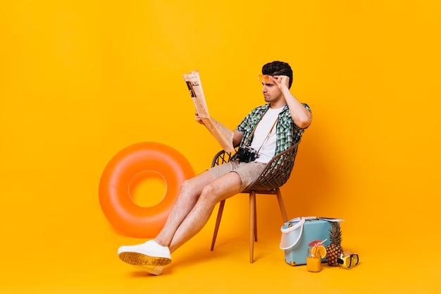 Free photo man in summer outfit removes glasses and reads newspaper. portrait of guy on orange space with suitcase and rubber ring.