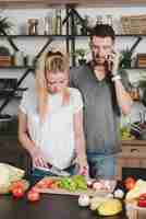 Free photo man talking on mobile phone looking at wife cutting vegetables with knife in the kitchen