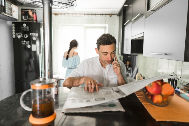 Free photo man talking on mobile phone reading newspaper with his wife standing at background