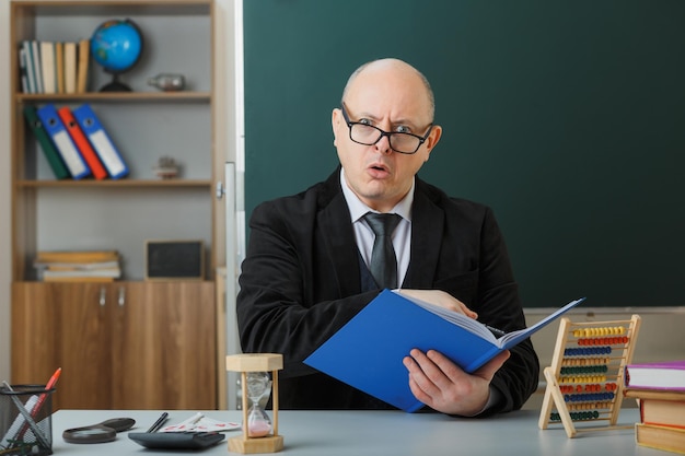 Free photo man teacher wearing glasses checking class register looking at camera being confused and surprised sitting at school desk in front of blackboard in classroom