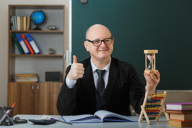 Free photo man teacher wearing glasses sitting at school desk in front of blackboard in classroom holding hour glass explaining lesson showing thumb up happy and pleased