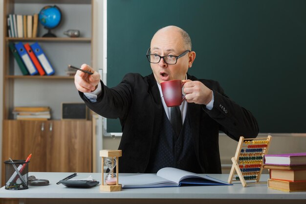 Man teacher wearing glasses sitting at school desk in front of blackboard in classroom holding mug of coffee pointing with pencil at something being surprised