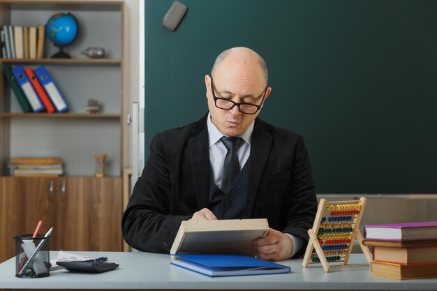 Man teacher wearing glasses sitting at school desk in front of blackboard in classroom preparing for lesson with serious face