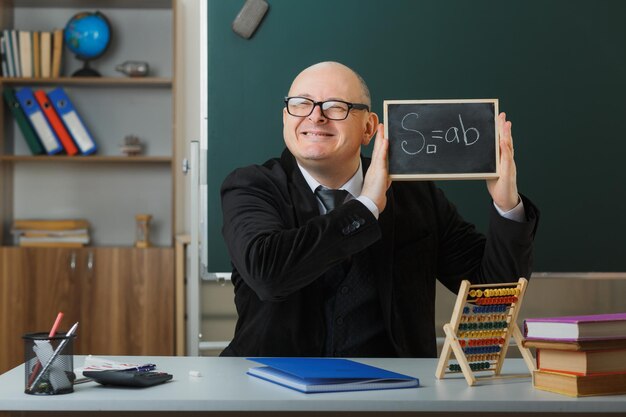 Man teacher wearing glasses sitting at school desk in front of blackboard in classroom showing chalkboard explaining lesson happy and pleased smiling cheerfully