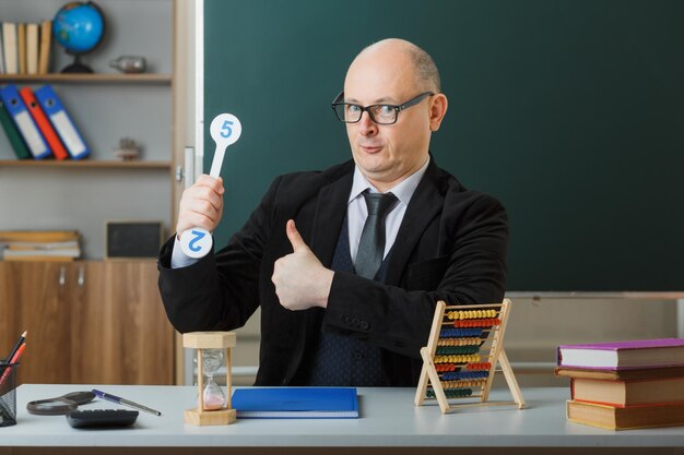 Man teacher wearing glasses sitting at school desk with class register in front of blackboard in classroom holding number plates explaining lesson showing thumb up happy and positive