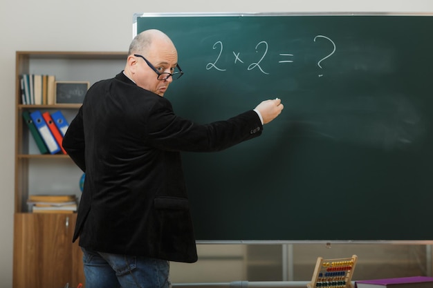 Man teacher wearing glasses standing near blackboard in classroom explaining lesson looking confused