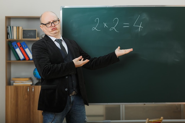 Man teacher wearing glasses standing near blackboard in classroom explaining lesson looking disappointed