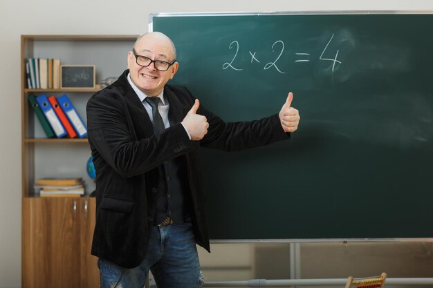 Man teacher wearing glasses standing near blackboard in classroom explaining lesson showing thumbs up happy and pleased