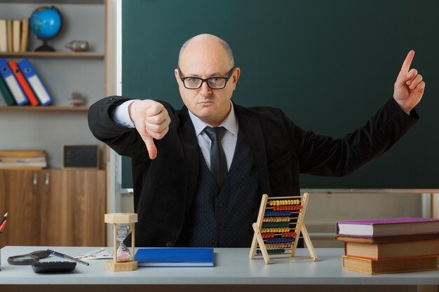 Man teacher wearing glasses with class register sitting at school desk in front of blackboard in classroom looking displeased showing thumb down pointing with index finger up