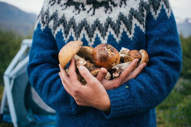 Free photo man in traditional blue wool sweater with ornaments stands on camping ground in mountains, holds in arms pile of delicious and organic, fresh natural mushrooms from forest