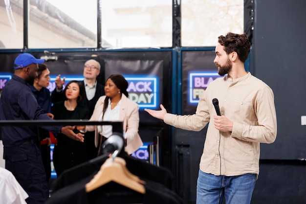 Free photo man tv reporter standing at shopping mall entrance presenting news about black friday. clothing store security guard opening door for eager people shoppers waiting for discounts and seasonal sales