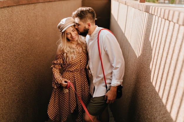 Free photo man in white shirt and khaki shorts gently kisses his positive smiling blonde wife in brown dress.