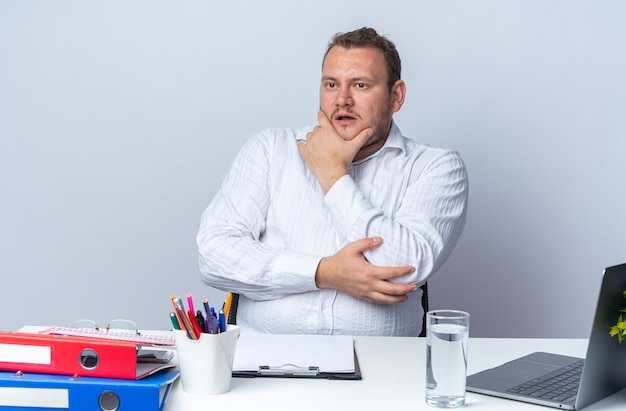 Free Photo man in white shirt looking aside with pensive expression with hand on his chin sitting at the table with laptop office folders and clipboard over white wall working in office