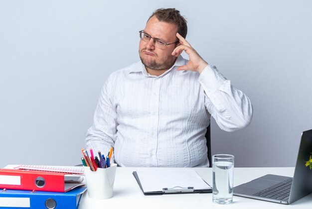 Man in white shirt wearing glasses looking aside confused and very anxious sitting at the table with laptop office folders and clipboard over white wall working in office