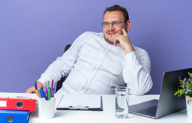 Man in white shirt wearing glasses looking aside smiling confident thinking positive sitting at the table with laptop and office folders over blue wall working in office