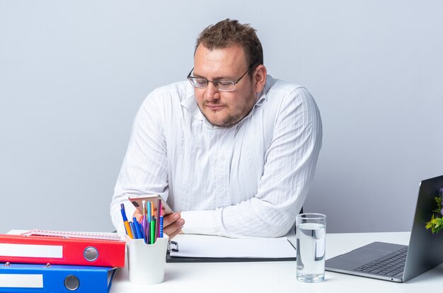 Man in white shirt wearing glasses looking tired and bored sitting at the table with laptop office folders and clipboard over white wall working in office