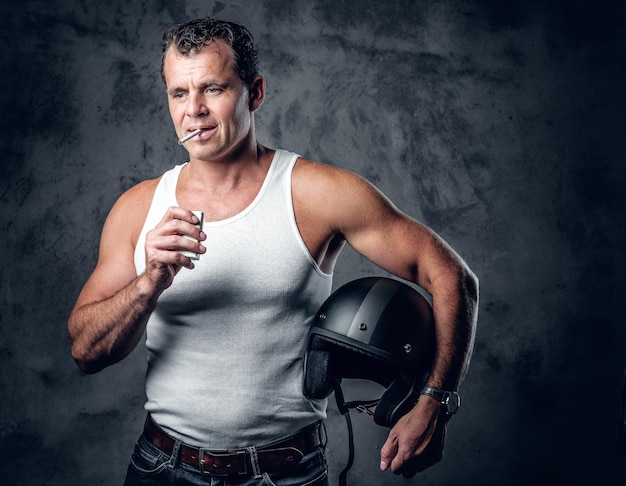 A man in a white t shirt, smoking cigarette and holds a motorcycle helmet in a photo studio.