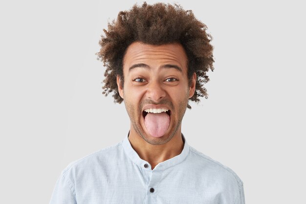 Man with Afro hairstyle shows tongue as notices something disgusting, makes grimace, demonstrates stubborn character