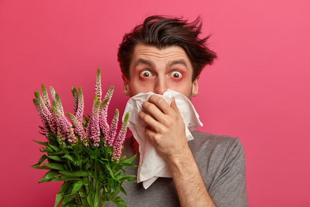 Free photo man with allergy sneezes and covers nose with napkin, listens advice from allergist how to cure hay fever, has red watery eyes, needs to treat allergic rhinitis, isolated on pink wall.