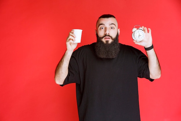 Free Photo man with beard holding an alarm clock and a cup of coffee.