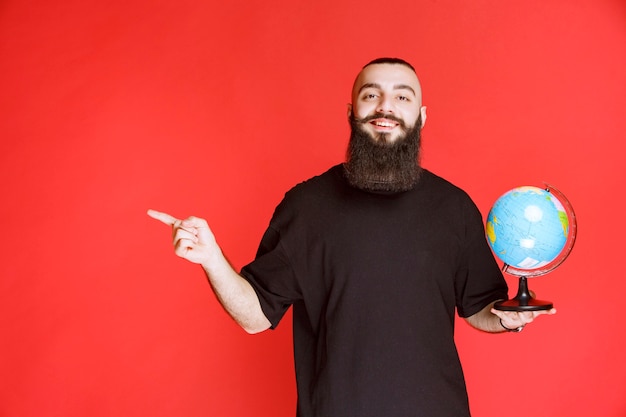 Free photo man with beard holding a globe and pointing to somewhere.