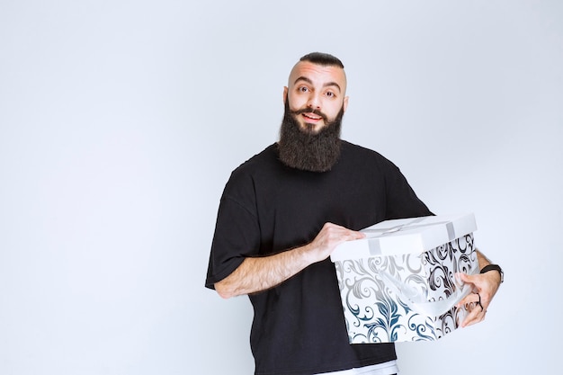 Free photo man with beard holding a white blue gift box and looks surprized.