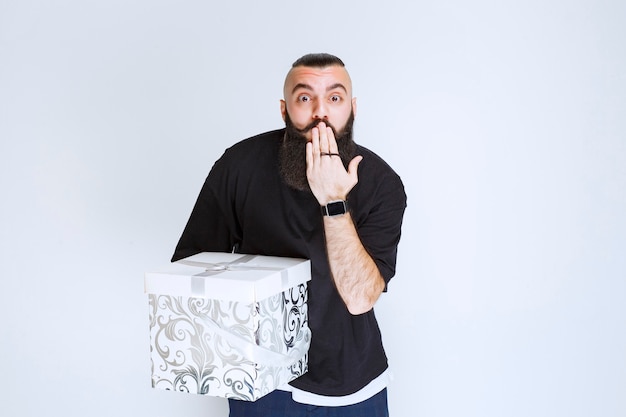 Free photo man with beard holding a white blue gift box pointing his mouth and asking for silence.