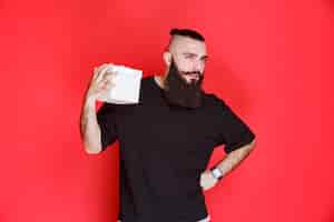 Free photo man with beard holding a white gift box and looks doubtful about what is inside.