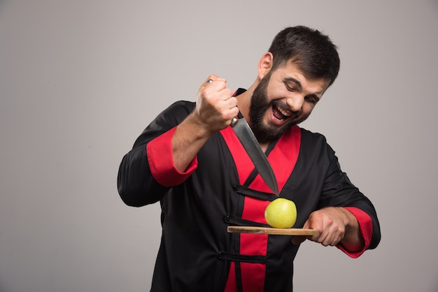 Man with beard trying to cut an apple on wooden board .